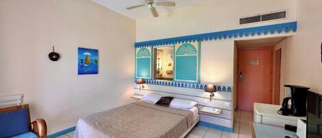 A hotel room with a bed, a desk, a chair, a ceiling fan, a painting, and a bathroom door.
