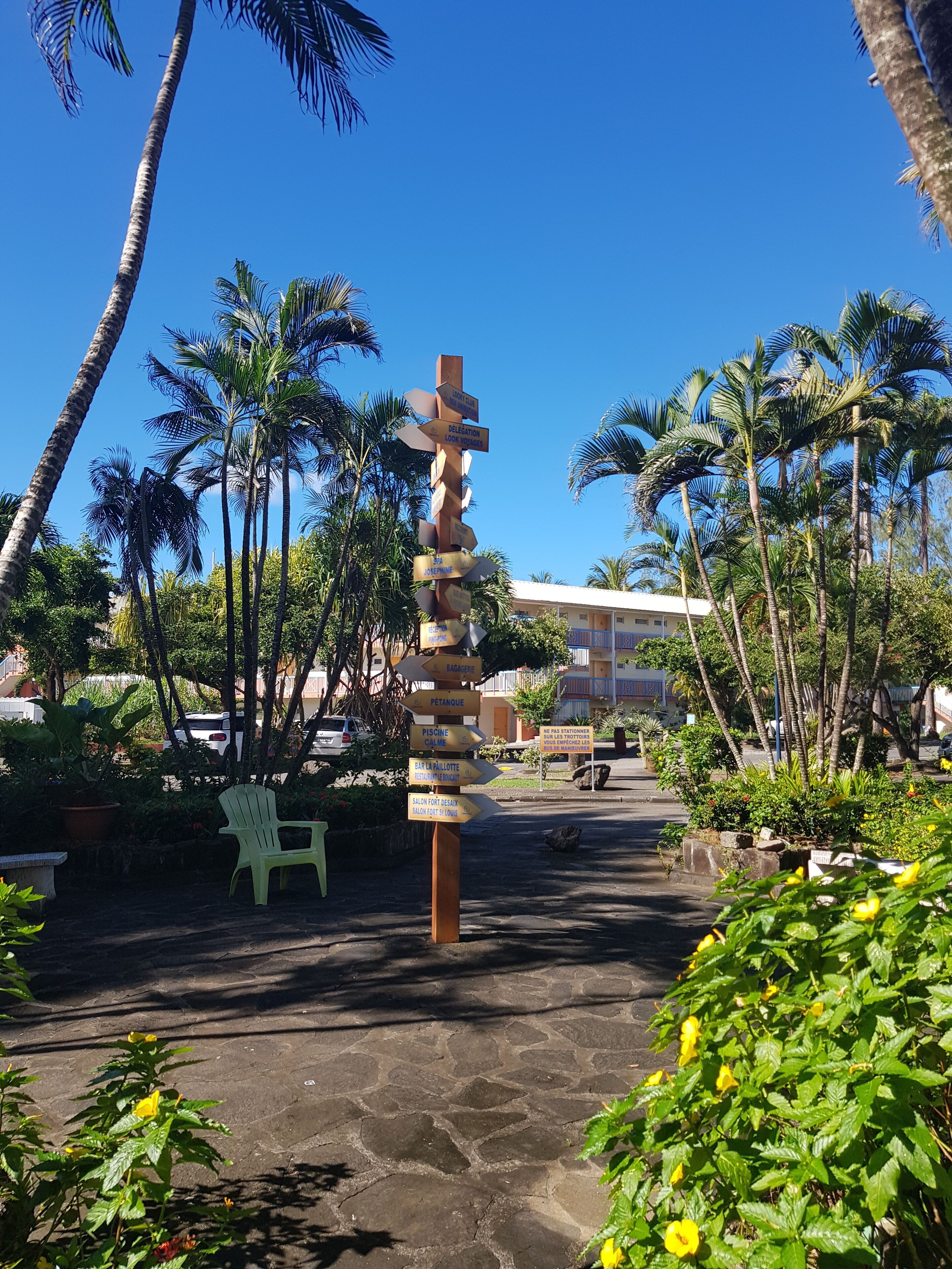 A wooden signpost with multiple directional arrows, surrounded by palm trees and a green chair in a sunny outdoor setting.