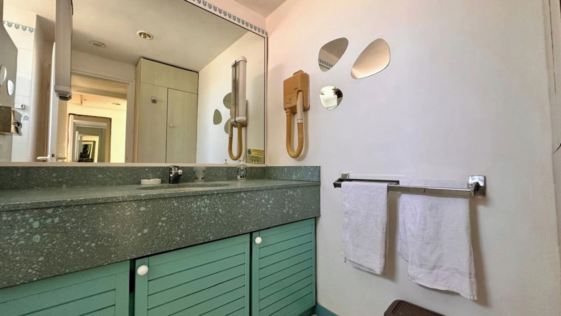 Uma casa de banho com bancada de granito, um armário verde, um espelho grande, um porta-toalhas com duas toalhas e um chuveiro com chuveiro manual.