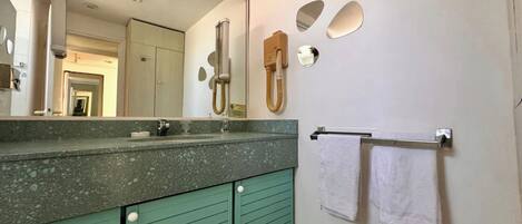 Uma casa de banho com bancada de granito, um armário verde, um espelho grande, um porta-toalhas com duas toalhas e um chuveiro com chuveiro manual.