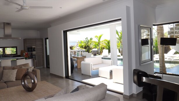 Ein modernes Wohnzimmer mit einer Couch, einem Couchtisch und einem Essbereich mit Tisch und Stühlen. Es gibt eine Küchenzeile mit einer Mikrowelle und einem Spülbecken. Der Raum verfügt über große Fenster und bietet Blick auf einen Pool und Grünflächen.