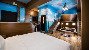Habitació d'hotel amb un llit gran, un escriptori, una cadira, una taula petita, un bany i un mirall. A la paret hi ha un mural que representa un cel i núvols.