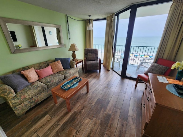 A living room with a sofa, armchair, and a table. There is a large mirror on the wall and a view of the beach through the glass door.