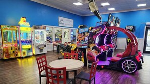 Ett spelhall med spelautomater där man kan fånga priser med en gripklo, ett racingspel och ett bord med stolar.