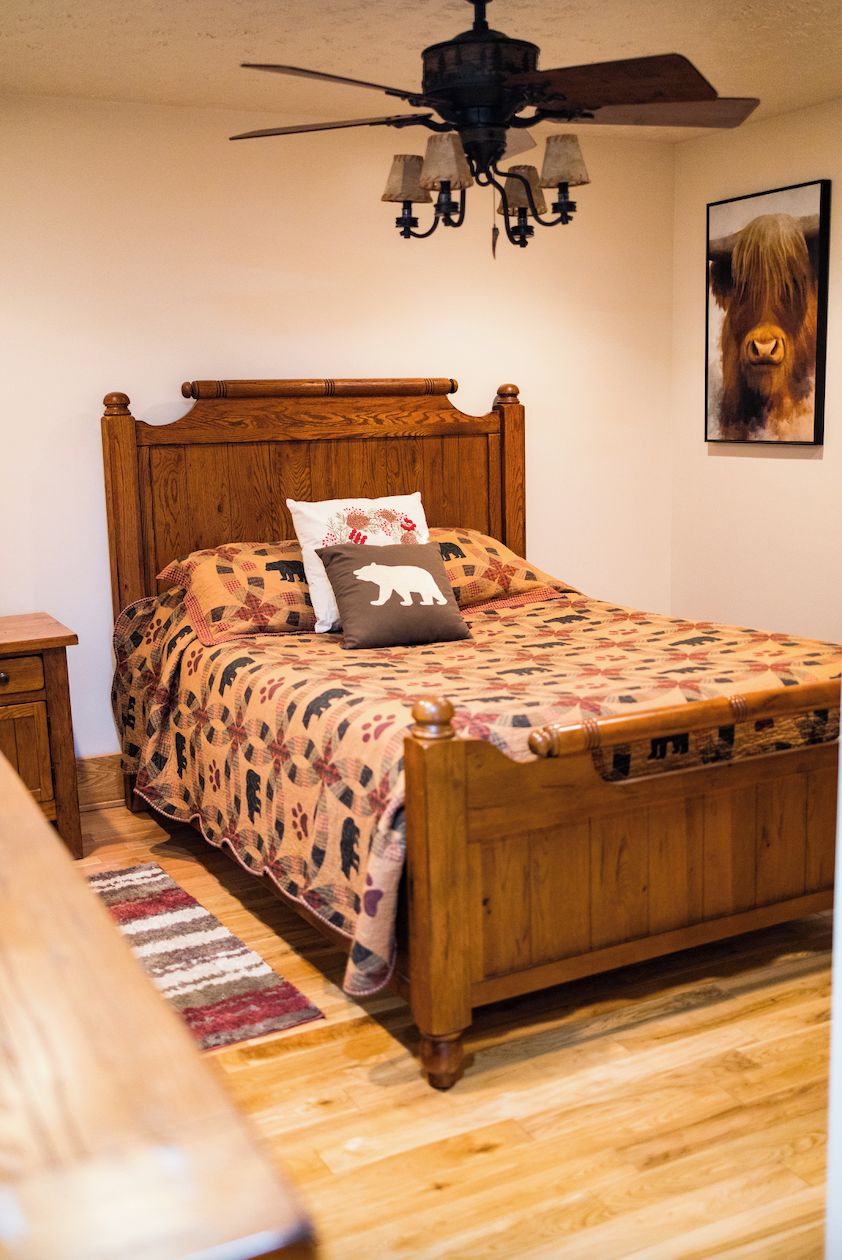 A wooden bed with a patterned bedspread and two pillows, a ceiling fan, and a rug on a wooden floor.