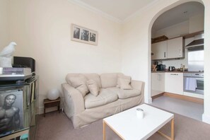 A living room with a beige sofa, a white coffee table, and a small side table with a lamp. A framed photo is on the wall, and a kitchen is visible through an archway.