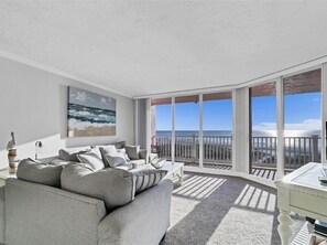 Un salón amplio con un sofá gris, un cuadro del océano y un balcón con vista al mar.