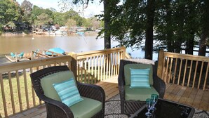 Una terraza de madera con sillas de mimbre y una mesa, con vista a un lago con barcos y casas al fondo.