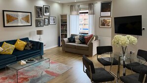 A modern living room with a blue sofa, a beige sofa, a glass coffee table, and a dining table with chairs. There are framed pictures on the walls, a bookshelf, and a flat-screen TV.