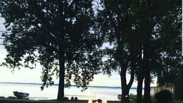 A fire pit with flames, surrounded by grass and trees, overlooking a body of water. - Your comfortable vacation place! (Plattsburgh)
