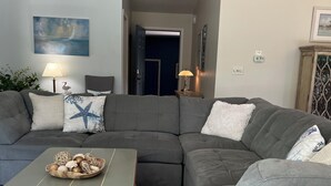 En stue med en hjørnesofa, et sofabord og dekorative puder. Et maleri hænger på væggen, og en lampe står på et sofabord.