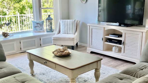 En stue med en beige sofa, en hvid lænestol, et sofabord med dekorative genstande, et fladskærms-tv på et træskab og et vindue med udsigt til grøn vegetation.