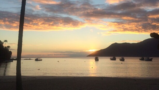 Un coucher de soleil sur une mer calme, avec des bateaux, des palmiers et une chaîne de montagnes en arrière-plan.