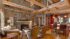 Amplia sala de estar con chimenea de piedra, vigas de madera y techo alto. Hay un rincón de estar acogedor con un sofá y sillones, y un comedor con mesa y sillas.