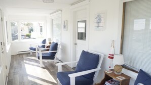 Um quarto claro e ensolarado, com paredes brancas e chão de madeira. Há duas poltronas brancas com almofadas azuis, uma pequena mesa de madeira com um abajur, e um quadro emoldurado na parede.