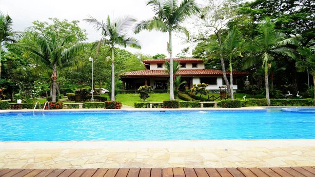 A swimming pool with a wooden deck in the foreground, surrounded by lush greenery and palm trees. In the background, there is a two-story building with a red-tiled roof and a statue in front. - Quinta El Poblado Luxury Home near Jaco Beach (Provincia de Alajuela)