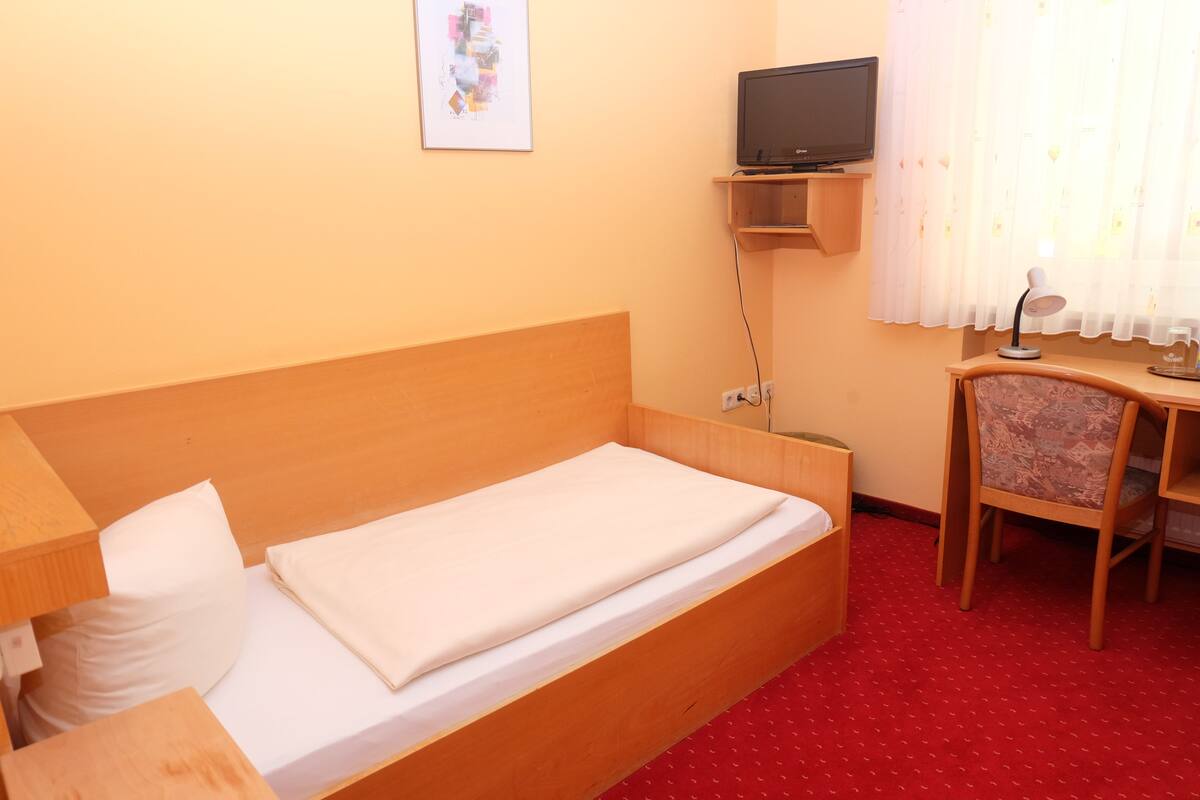 A single bed with a wooden headboard, a bedside table, a chair, a TV on a shelf, and a window with curtains.
