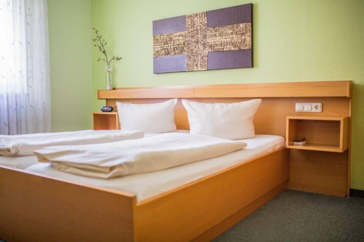 A neatly made bed with white linens, a wooden headboard, a bedside table with a lamp, and a wall-mounted artwork.