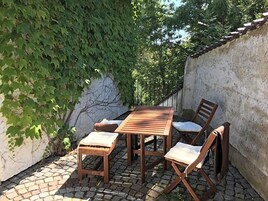 Un juego de mesa y sillas de madera sobre un patio adoquinado, con una pared blanca y follaje en el fondo.