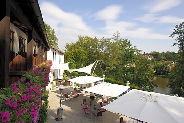 An outdoor seating area with tables and umbrellas, overlooking a river and greenery.
