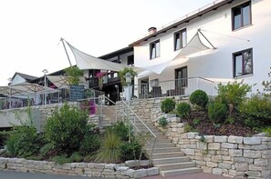 A building with a stone staircase leading to a covered entrance, surrounded by greenery and a white awning.