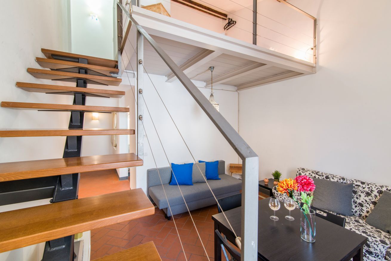 A modern interior with a wooden staircase, a grey sofa with blue cushions, a black table with two glasses and a vase of flowers, and a patterned sofa.