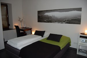 A hotel room with a bed, a desk, a chair, a nightstand, and a wall-mounted picture.