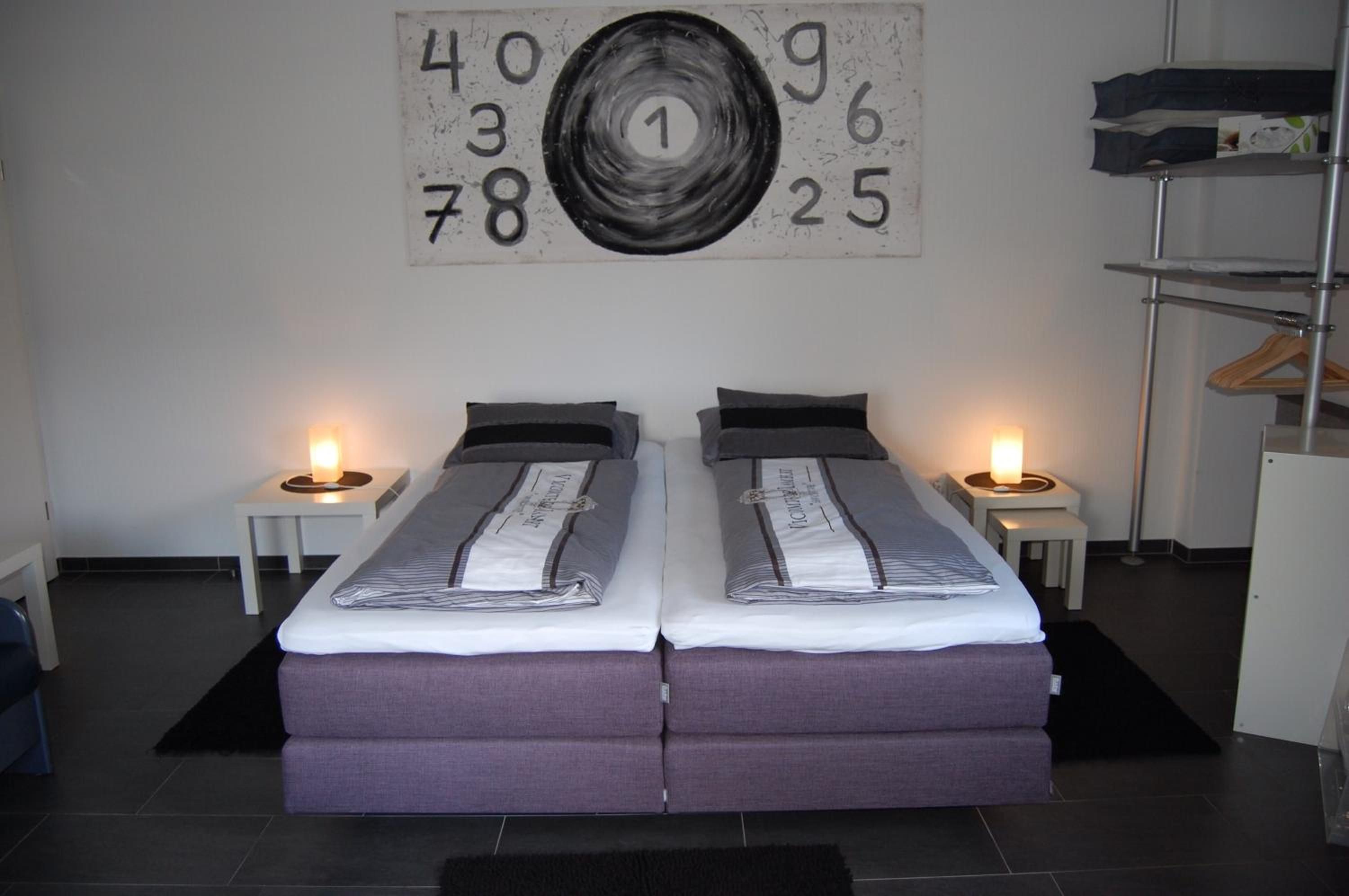 A double bed with a grey and white striped duvet, two bedside tables with lamps, and a wall with a large abstract number painting.