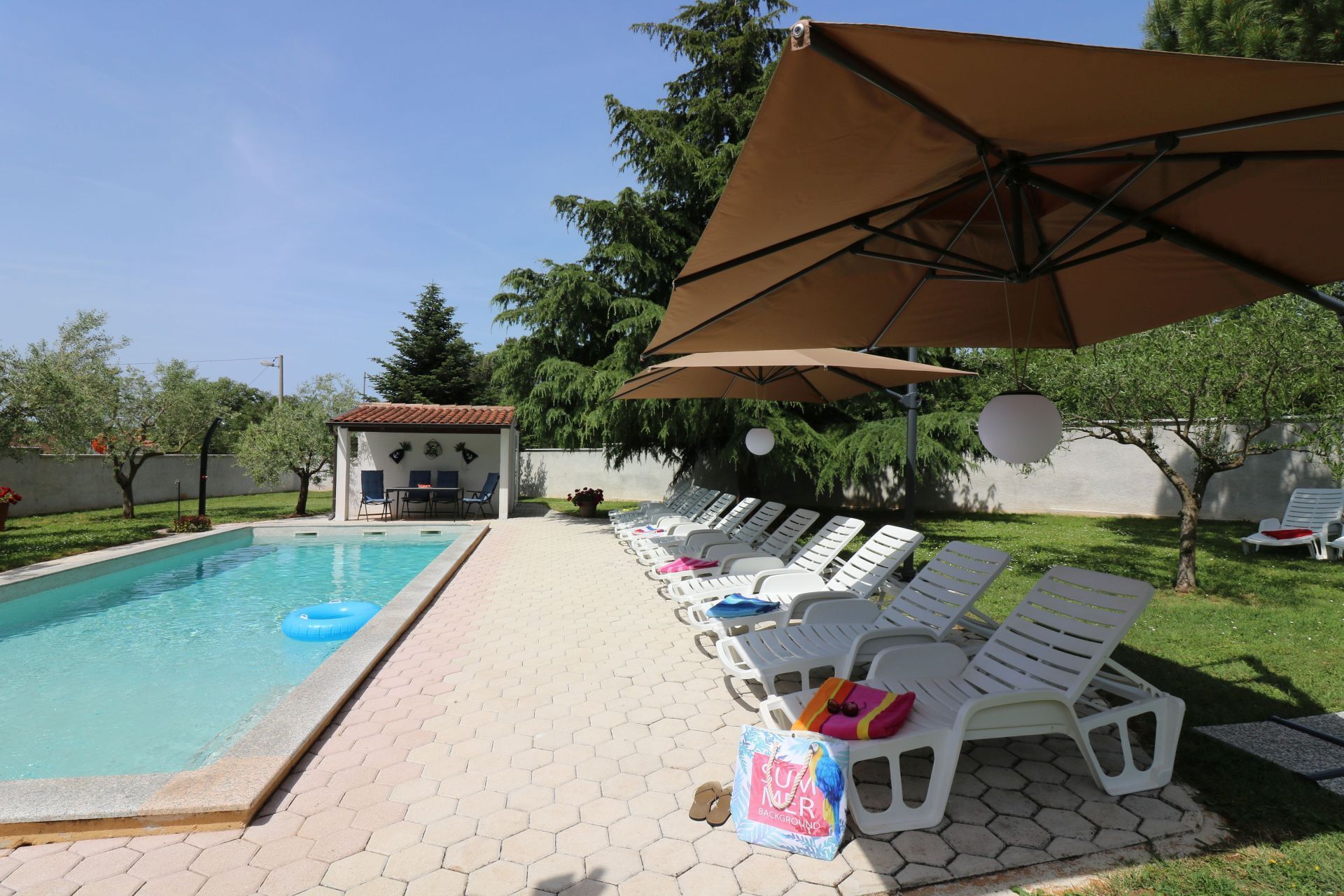Una piscina con sillas de descanso y sombrillas, un pequeño pabellón y un área verde con árboles.