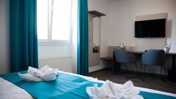 A hotel room with a bed, a desk, two chairs, a flat-screen TV, and a window with curtains.
