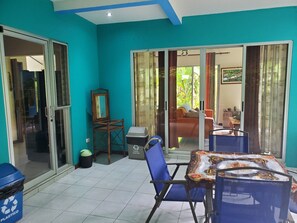 Une pièce avec un mur bleu, une table recouverte d’une nappe à motifs, une chaise, une poubelle et une porte donnant sur un espace extérieur aménagé avec un canapé et des rideaux.