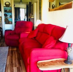 A red sofa in a room with a wooden floor, a lamp on a side table, and shelves with various items.
