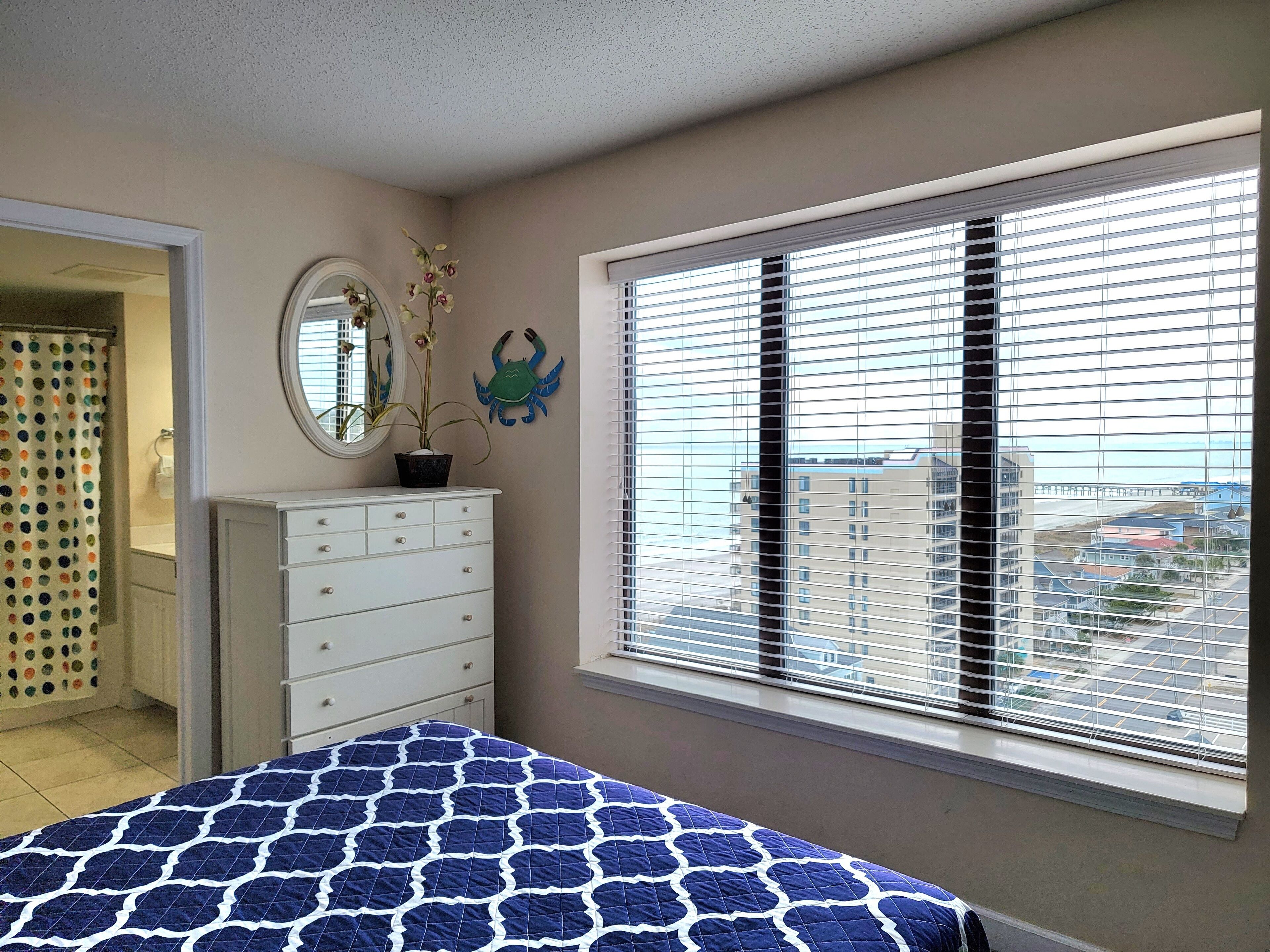 A bedroom with a bed, a dresser, a mirror, a window with blinds, and a view of buildings.