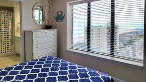 Quarto com cama, cómoda, espelho, janela com persianas e vista para edifícios.