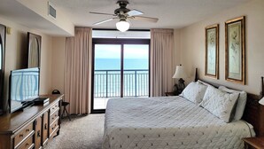 Quarto de hotel com uma cama grande, um ventilador de teto, televisão, cômoda e vista para o oceano.
