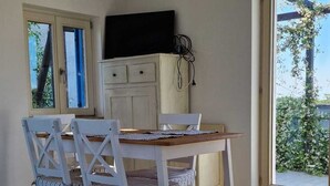 Ruokailutilassa on puinen pöytä ja tuolit, televisiotaulutaso ja suuri ikkuna, josta avautuu näkymä vehreään maisemaan.