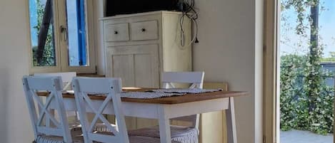A dining area with a wooden table and chairs, a television on a cabinet, and a large window with a view of greenery.