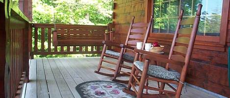 A wooden porch with rocking chairs and a table, surrounded by a wooden fence and a window with blinds.