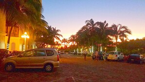 Ein gepflasterter Bereich mit geparkten Autos und Palmen, ein Gebäude mit Säulen und ein Sonnenuntergang im Hintergrund.