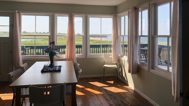Ein Essbereich mit Tisch und Stühlen, großen Fenstern mit Vorhängen sowie Blick auf Strand und Felder.