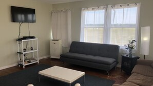 Ein Wohnzimmer mit grauem Sofa, einem weißen Couchtisch, einem an der Wand montierten Fernseher und einem Fenster mit Vorhängen.