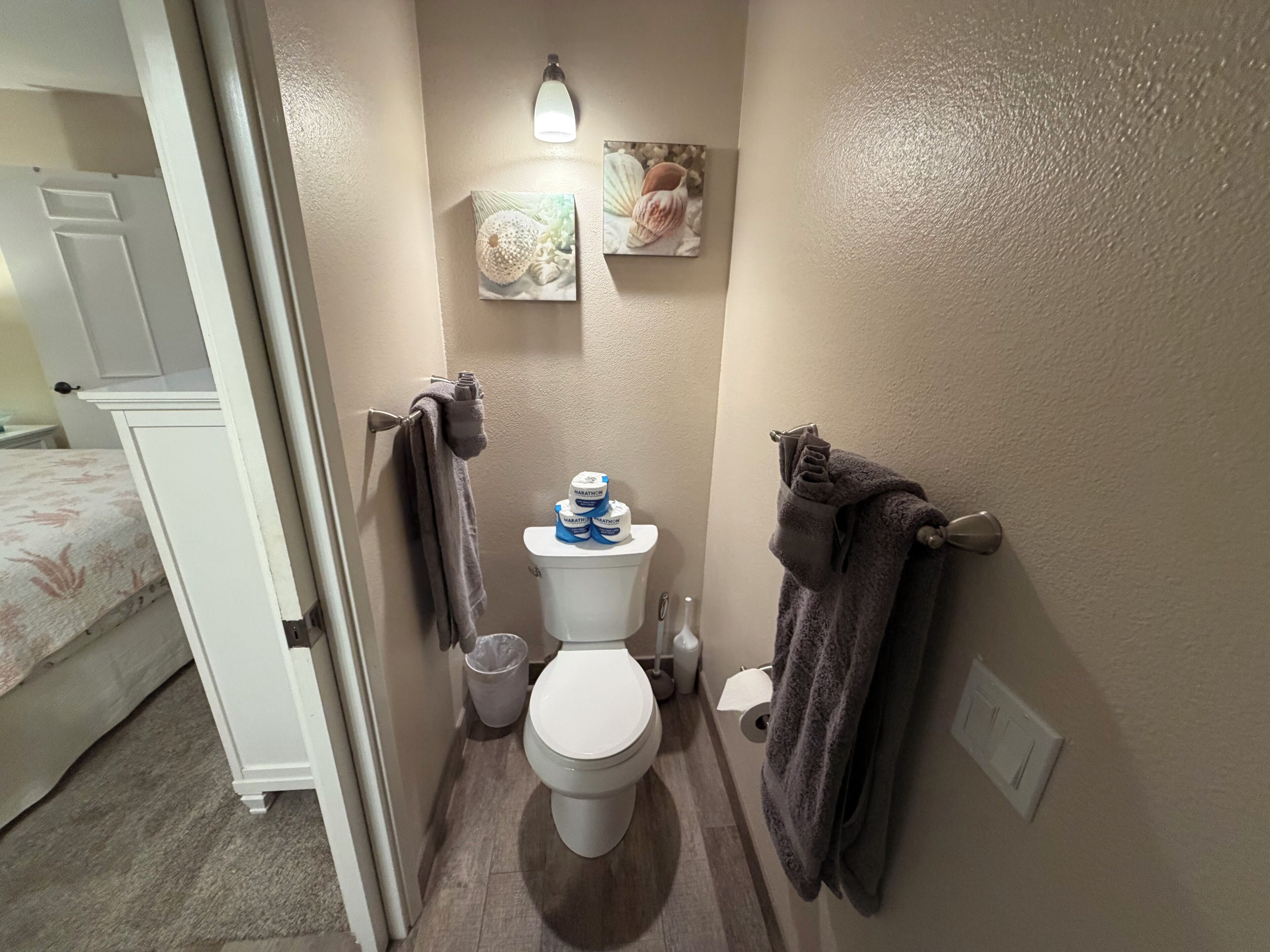 Une petite salle de bain avec des toilettes, un lavabo et une douche. Des serviettes sont accrochées au mur et un luminaire est fixé au-dessus des toilettes.