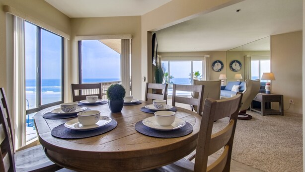 A dining area with a wooden table set for four, large windows offering a view of the ocean, and a living area with a sofa and a lamp. - 5 - STAR -⭐⭐⭐⭐⭐- LUXURY OCEAN FRONT CONDO - JACUZZI - FAMILY FRIENDLY - SLEEPS 6 (San Diego)