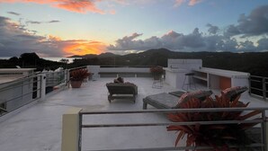 Una terraza en la azotea con mobiliario exterior, plantas en macetas y vistas al atardecer sobre un paisaje montañoso.