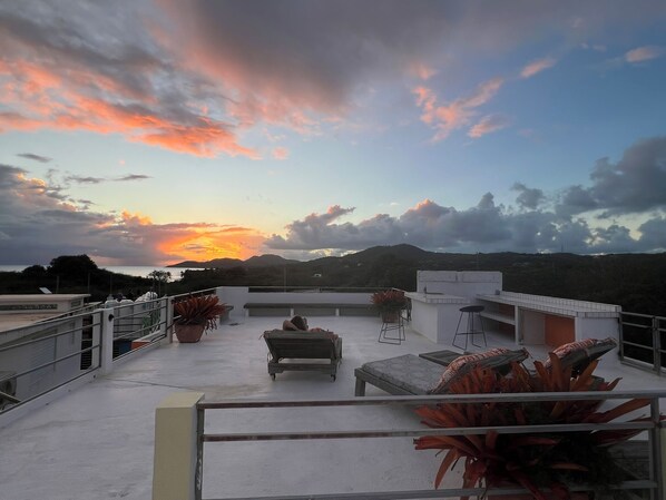 A rooftop terrace with outdoor seating, potted plants, and a view of the sunset over a mountainous landscape.