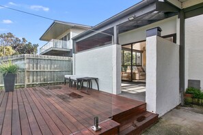 A wooden deck with a table and chairs, a white exterior wall, and a visible window.