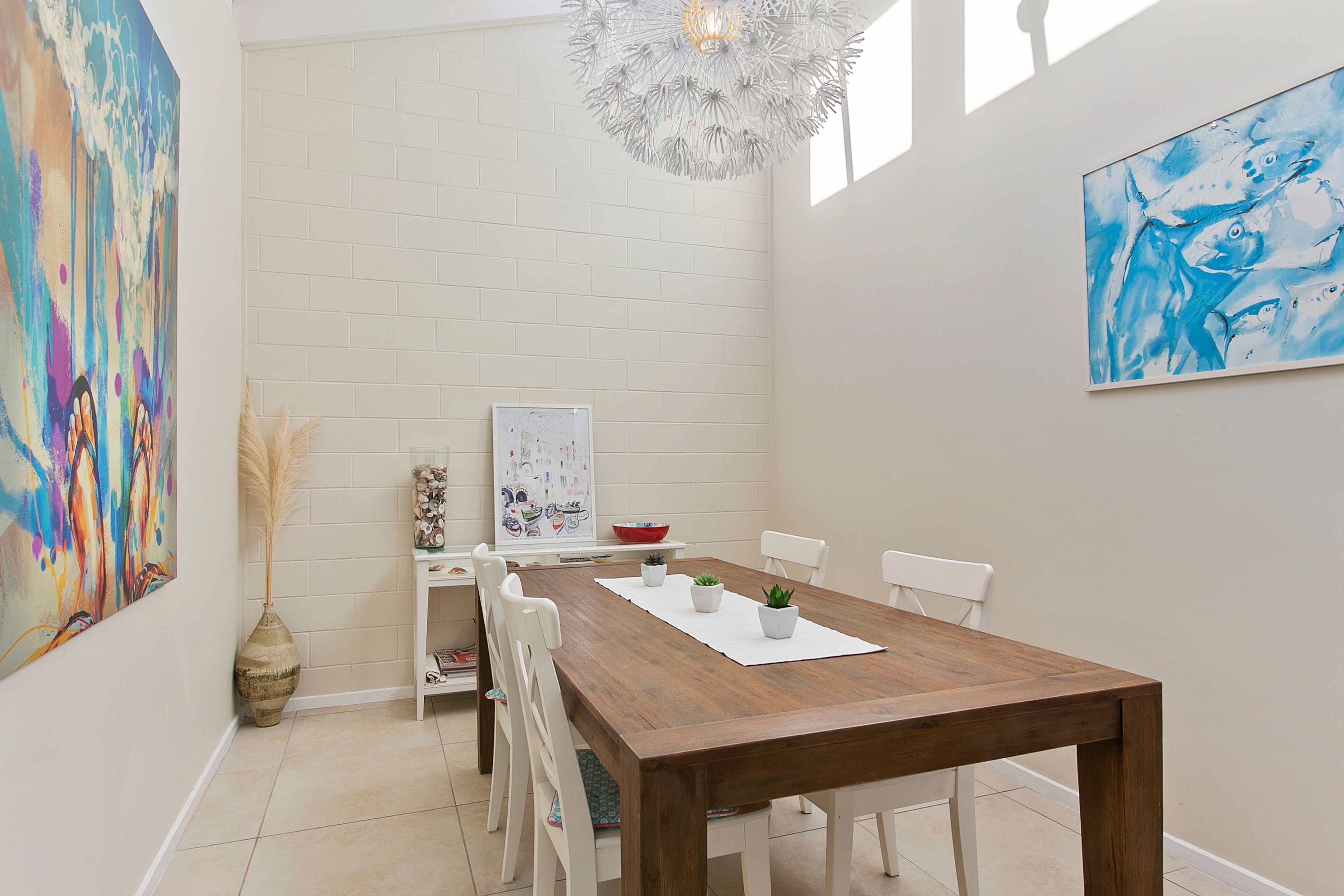 Un espace repas comprenant une table en bois, des chaises blanches, un mur en briques blanches et deux œuvres d’art.