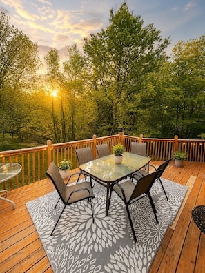 Una terraza con una mesa y sillas de vidrio, rodeada por una barandilla de madera, con vista a árboles y un atardecer.