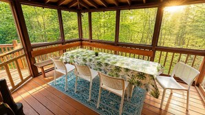 A covered outdoor dining area with a table, chairs, and a view of a forest.