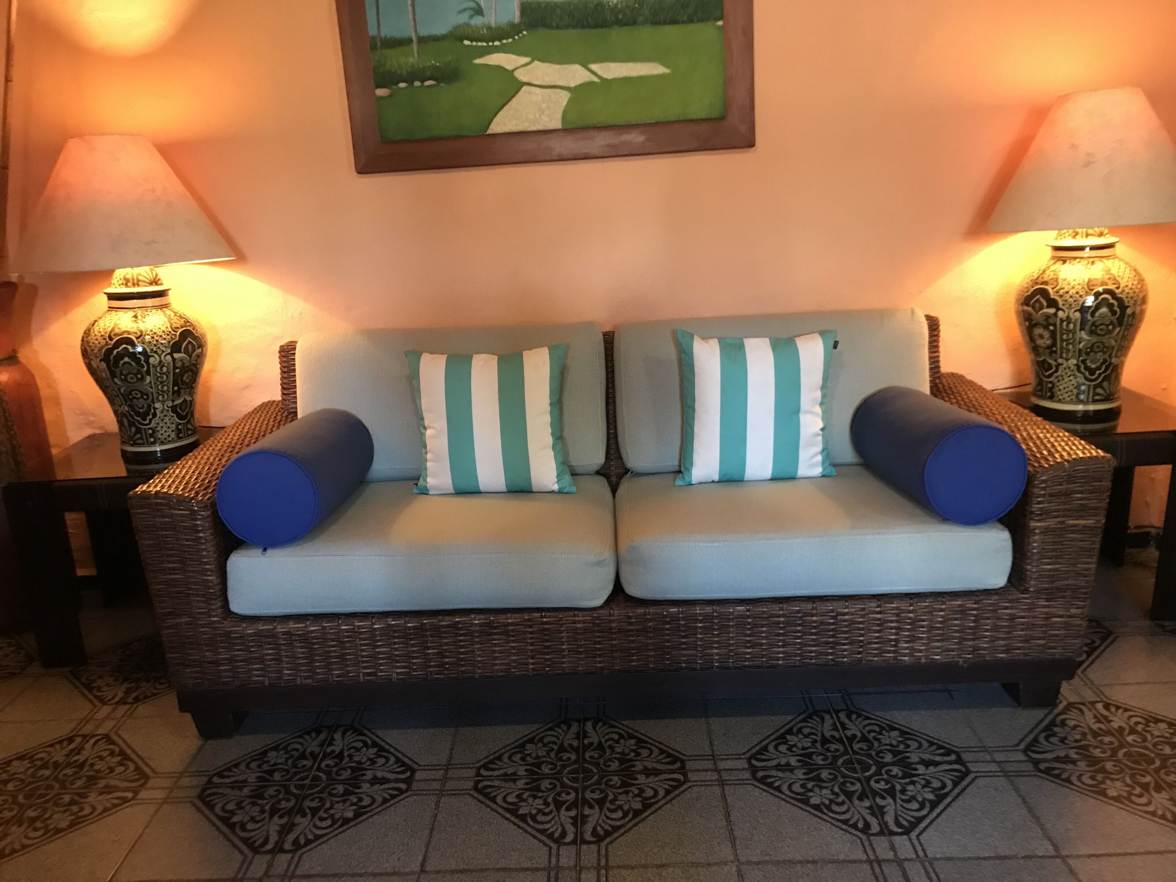 A wicker sofa with striped cushions and blue accent pillows, flanked by two ornate vases on matching side tables, with a framed landscape painting above.